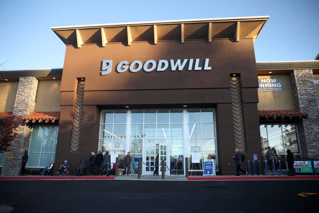 People wait in line outside a Goodwill store with a "Now Hiring" sign on the building and clear skies above.
