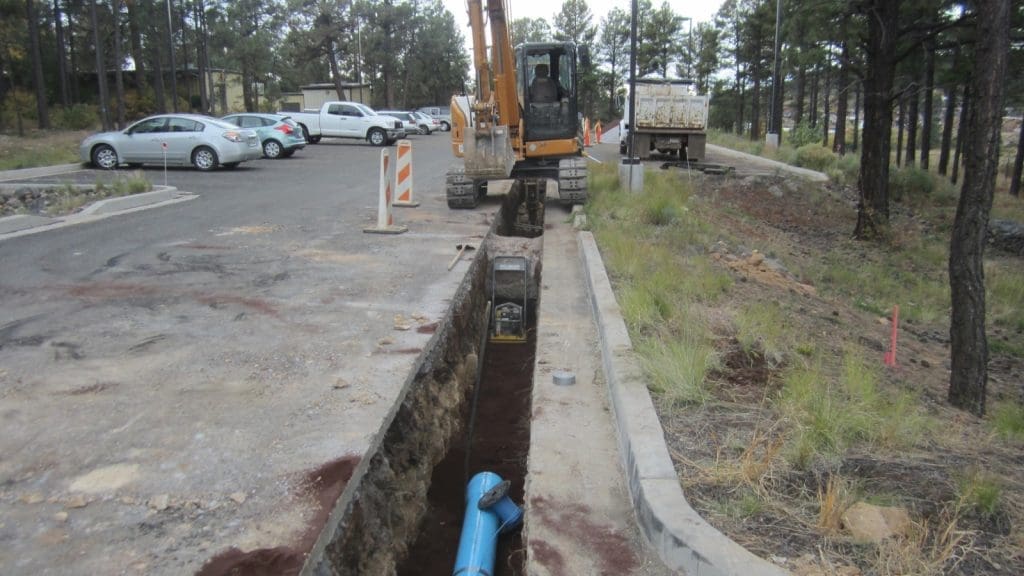 An excavator works beside a parking lot, digging a trench with a blue pipe partially installed underground. Traffic barriers and parked cars are visible in the background.