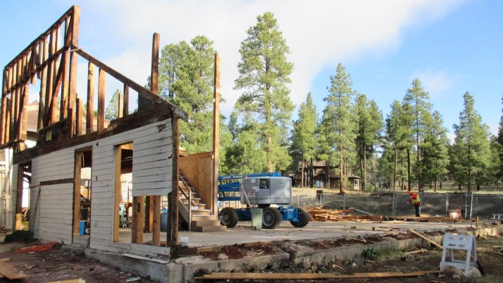 Partially demolished wooden building with exposed framework, construction equipment, and workers on site; pine trees and clear sky in the background.