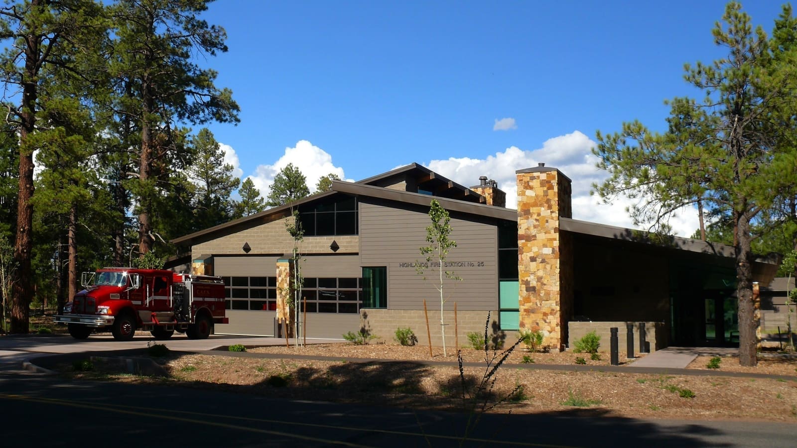A red fire truck is parked outside Highlands Fire Station No. 25, which recently received a design award, surrounded by tall pine trees under a clear blue sky.