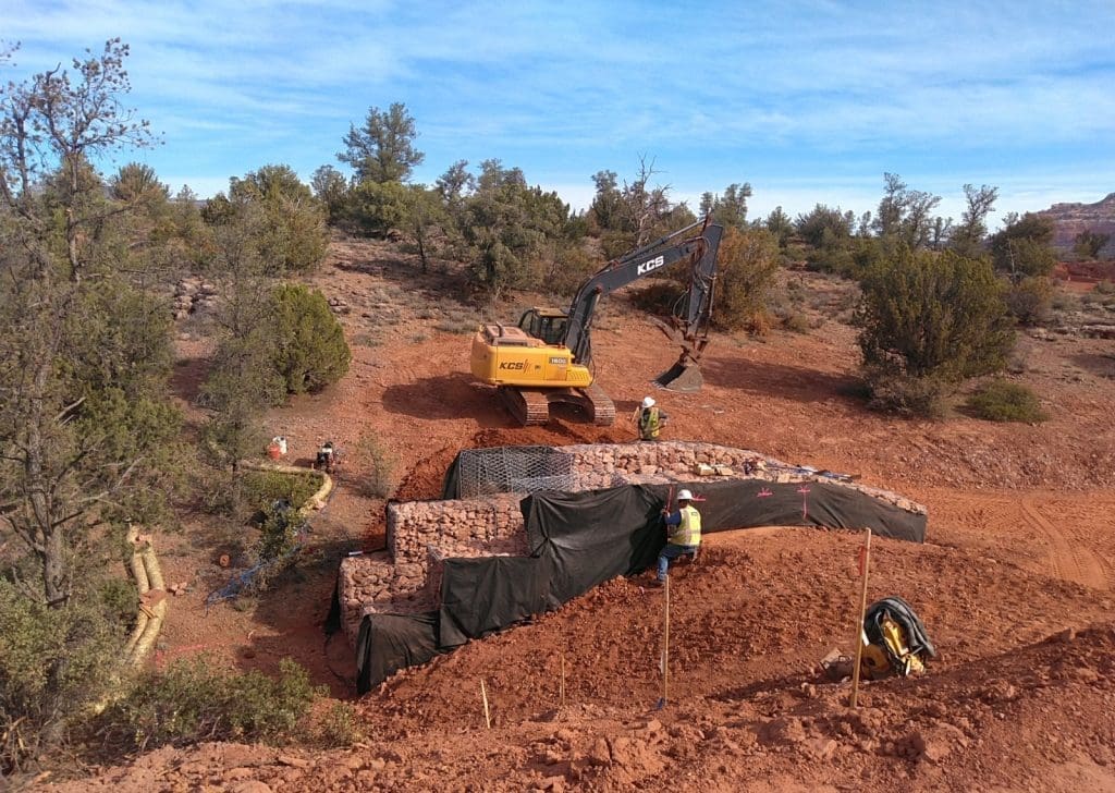 A construction site in a desert area shows workers and an excavator building a rock-filled retaining wall with a black protective barrier.
