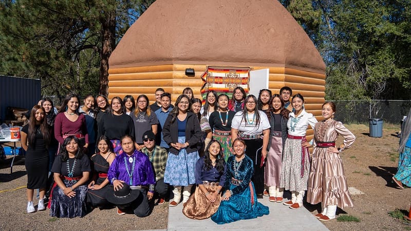 A group of young people in traditional and contemporary clothing pose in front of a log cabin structure on a sunny day.