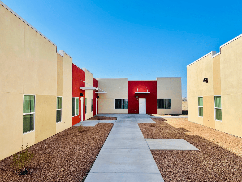 A modern, single-story building with red and beige walls and a concrete walkway, set against a clear blue sky and surrounded by gravel landscaping.