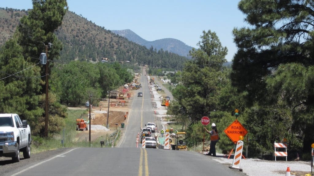 A rural road under construction with traffic cones, construction vehicles, and signs indicating "STOP" and "ROAD NARROWS." Trees and mountains are visible in the background.