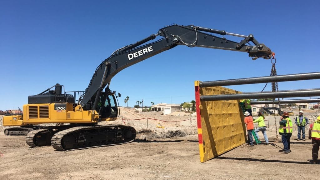 A large excavator lifts and positions a yellow trench shield while several construction workers in safety vests and helmets guide it at a worksite.