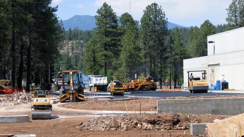 Construction site with heavy machinery and workers paving a road near a white building, surrounded by trees and mountains in the background.