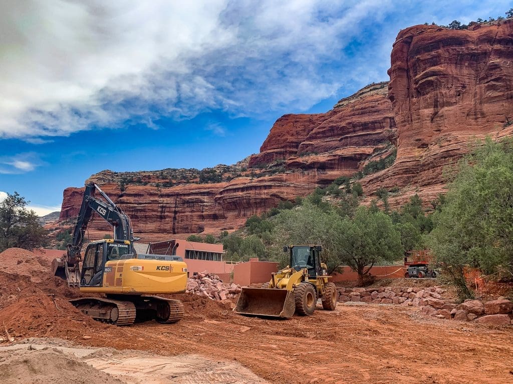 Excavator and front loader move red dirt at a construction site surrounded by red rock cliffs and scattered trees under a partly cloudy sky.