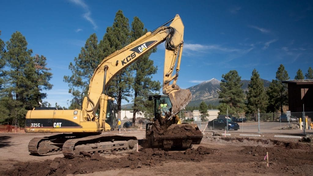 A yellow excavator operated by a worker is digging and moving soil at a construction site surrounded by trees and mountains.