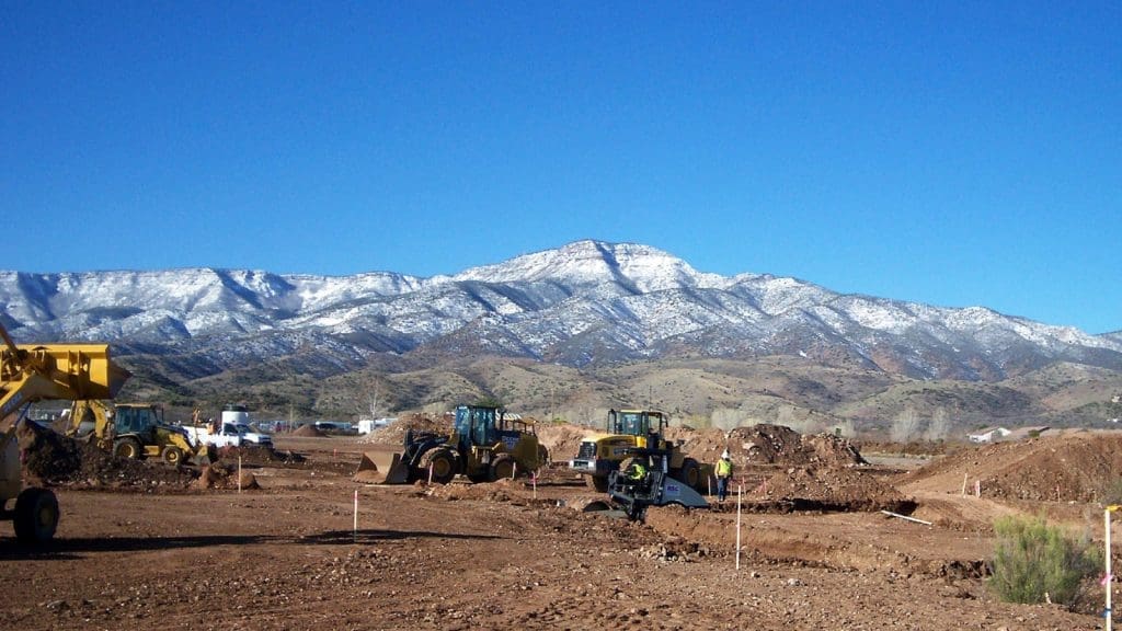 Construction vehicles and workers operate on a dirt site with a snow-capped mountain range in the background under a clear blue sky.
