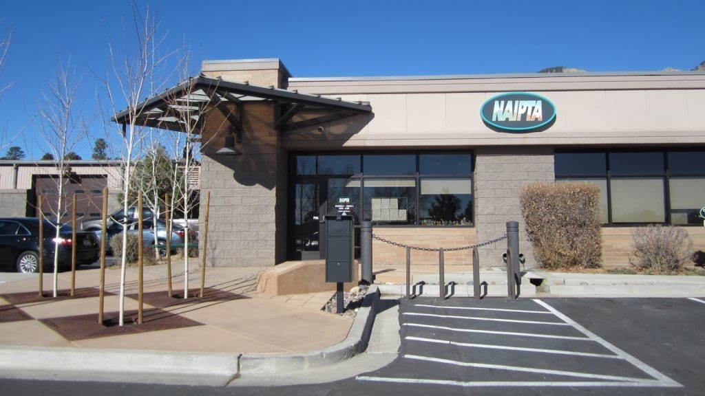 Single-story NAIPTA office building with large windows, a small awning over the entrance, a few leafless trees, and an accessible parking space in front.