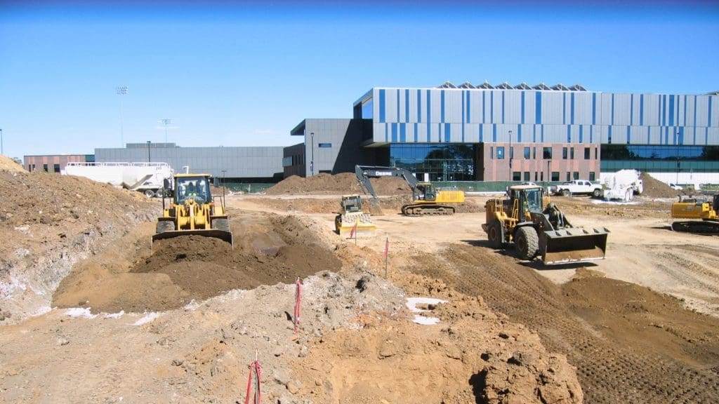 Construction site with several bulldozers and excavators moving earth in front of a modern building under a clear blue sky.