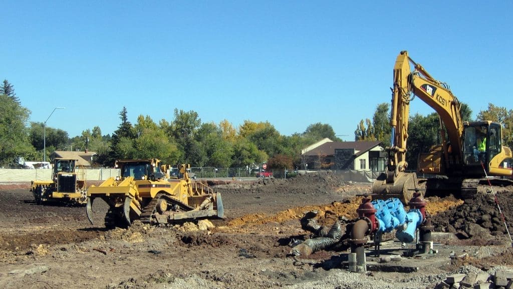 Construction site with two bulldozers and an excavator moving earth, near a residential area with trees and houses in the background.