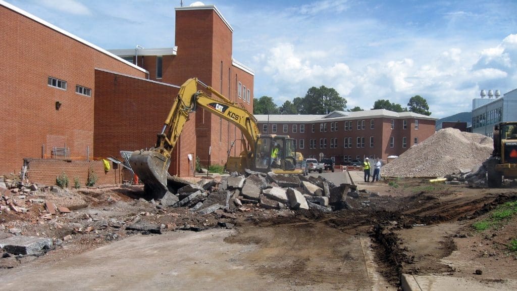 An excavator breaks up concrete outside a brick building while two workers stand nearby; piles of debris and dirt are visible on the construction site.