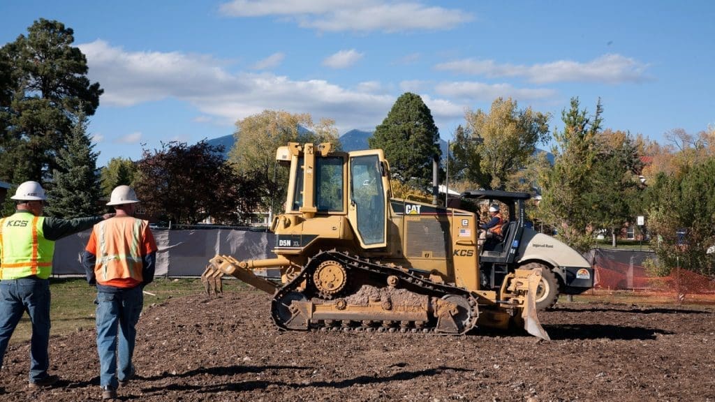 Two construction workers wearing safety gear stand near a yellow bulldozer and a roller on a dirt lot with trees and fencing in the background.