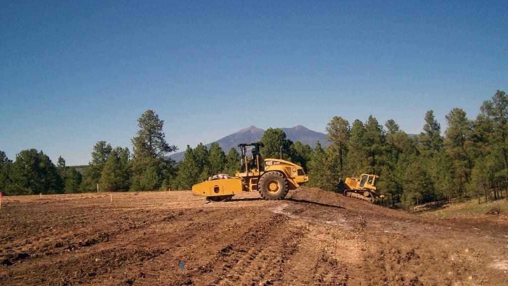 A yellow soil compactor and bulldozer work on a cleared dirt area surrounded by pine trees with mountains in the background under a clear blue sky.