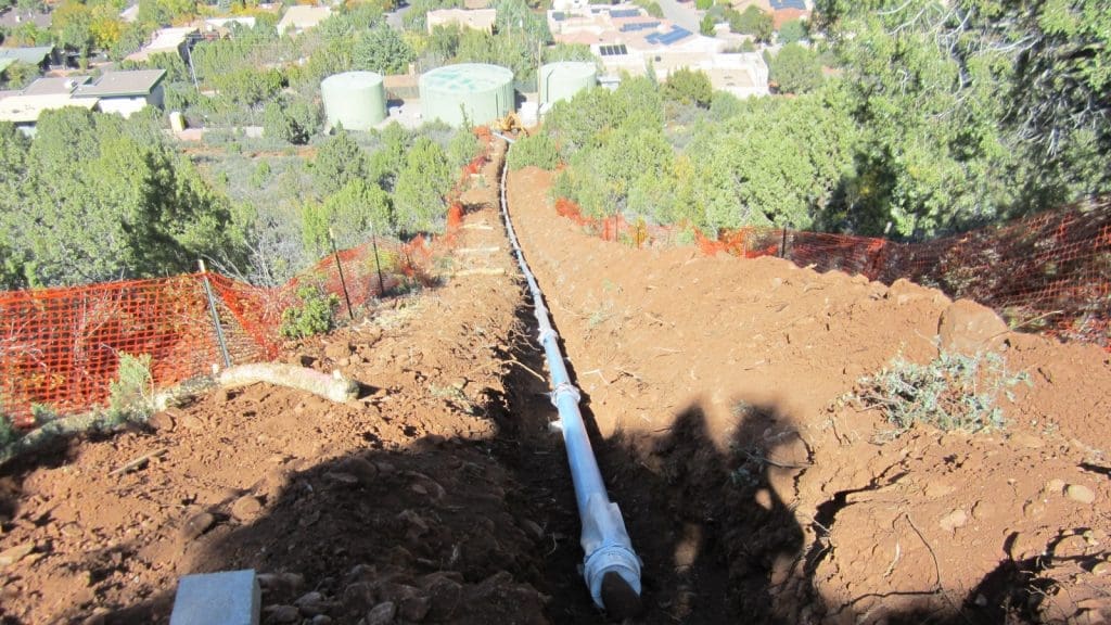 A pipeline installation runs down a dirt slope with orange safety fencing on both sides; storage tanks and buildings are visible in the background.