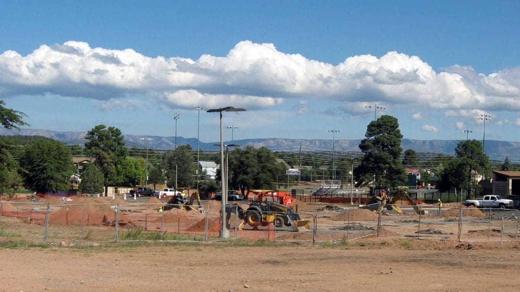 A construction site with several vehicles and equipment on dirt ground, surrounded by fencing, with trees and mountains in the background.