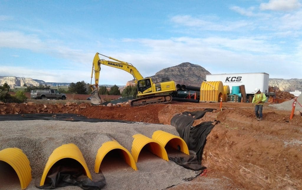 A yellow excavator moves earth at a construction site with exposed yellow plastic culverts, a worker in a safety vest, and hills in the background.