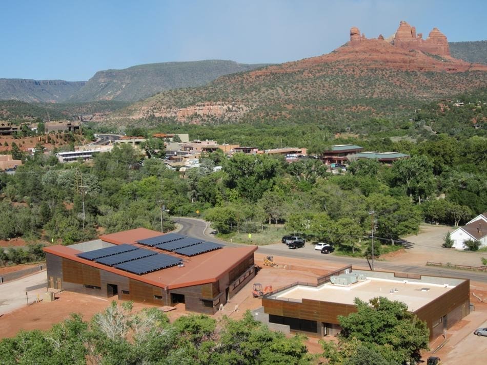 Aerial view of buildings, including the Sedona-Oak Creek USD District Office PV System, with solar panels nestled among green trees and hills, framed by striking red rock formations in the background.