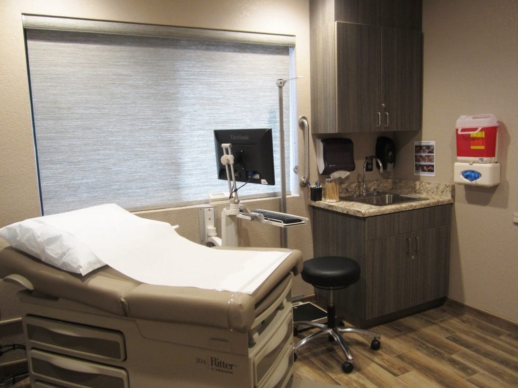 A medical exam room with an exam table, computer on a movable arm, sink, cabinets, stool, and medical supplies on the wall.