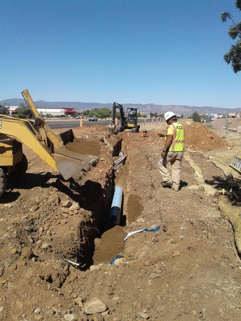 A construction worker observes an excavator and bulldozer digging a trench and laying a blue water pipe at a roadside construction site.