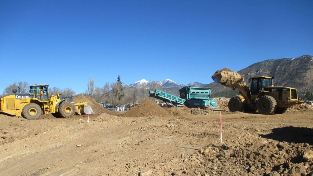 Two yellow bulldozers move dirt at a construction site with piles of soil, machinery, and mountains visible in the background under a clear blue sky.