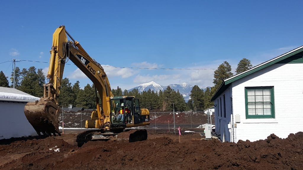 A yellow excavator is digging soil next to white buildings, with trees and snow-capped mountains visible in the background under a clear sky.