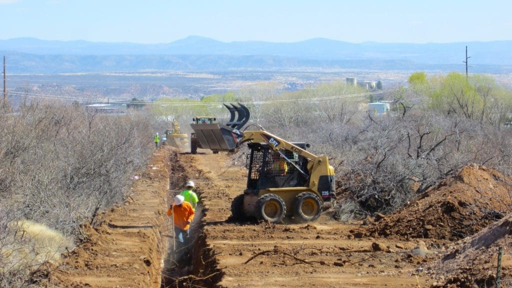 Construction workers operate heavy machinery and dig a trench along a dirt path surrounded by brush, with mountains visible in the background.