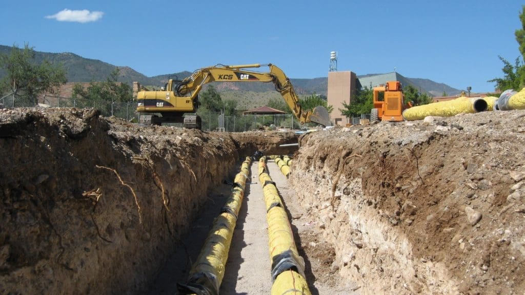 Excavator and heavy equipment at a construction site with yellow pipes laid in a long trench under clear blue sky.