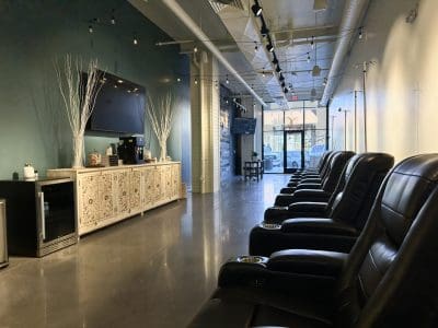 A row of black leather recliners lines a shiny floor in a modern lounge, with a long cabinet, TV, and decor along one wall; large windows provide natural light.