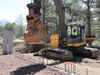 A construction worker operates a large excavator near a partially built concrete wall and rebar at a forested outdoor site.