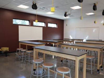 A classroom in the Northland Preparatory Academy STEAM Building features tall tables and stools, maroon and white walls, large windows, ceiling-mounted equipment, and two whiteboards.