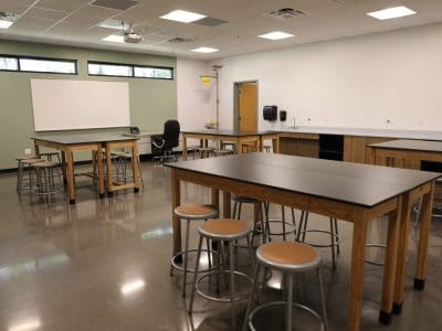 A modern science classroom in the Northland Preparatory Academy STEAM Building features lab tables, stools, a teacher’s desk, sinks, cabinets, and large windows. The room is bright and empty.