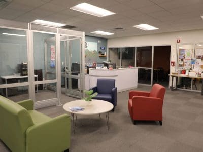 Office reception area with colorful chairs around a round table, a white reception desk, glass-walled office, and bulletin boards on the wall.