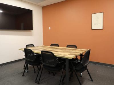 A small meeting room with a wooden table, six black chairs, a wall-mounted TV, orange accent wall, and a framed poster.