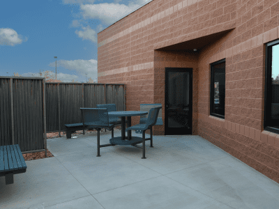 Outdoor patio area with a round table, three chairs, a bench, and a brick building wall under a partly cloudy sky.