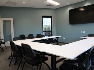 A conference room with a U-shaped arrangement of white tables, black chairs, a large wall-mounted screen, and a closed door leading outside.