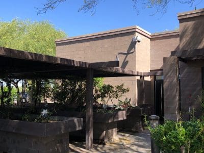 A modern brown brick building with a covered walkway, surrounded by greenery and trees under a clear blue sky.