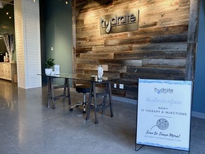 A modern reception area at Hydrate Bar with a wooden accent wall, glass desk, chair, plant, and informational sign about NAD+ IV therapy and injections.