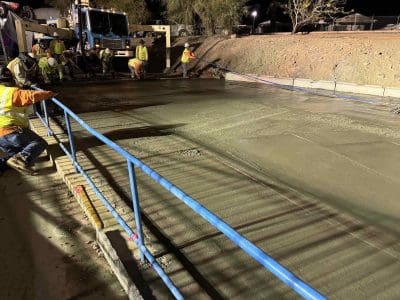 Construction workers pour and smooth concrete at a worksite at night, illuminated by artificial lights, with a blue handrail in the foreground.