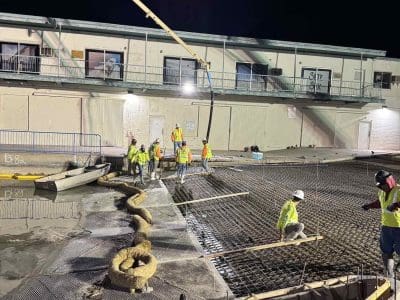 Construction workers pour and level concrete at night using a large concrete pump in front of a building. Erosion control wattles and reinforcement mesh are visible.