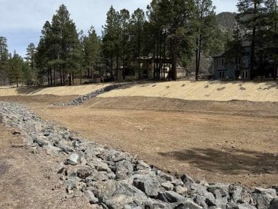 A dry, rocky drainage channel runs through a grassy area with pine trees and houses in the background under a partly cloudy sky.