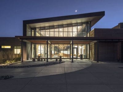 Modern building with large glass windows and angular roof, illuminated at dusk with outdoor seating and landscaped pathway in front.