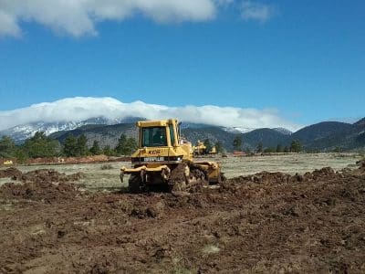 A yellow bulldozer moves earth on a muddy field with mountains and trees in the background under a partly cloudy sky.