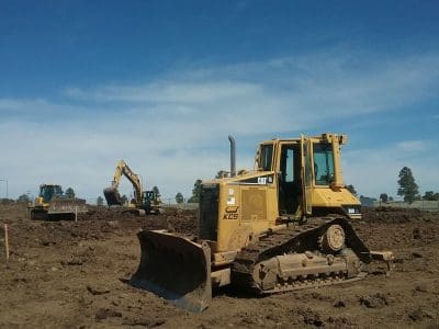Bulldozers and an excavator operate on a large construction site with dirt ground under a clear blue sky.