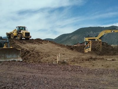 Three large construction vehicles, including a bulldozer, excavator, and loader, work on a dirt mound at an outdoor construction site with mountains in the background.