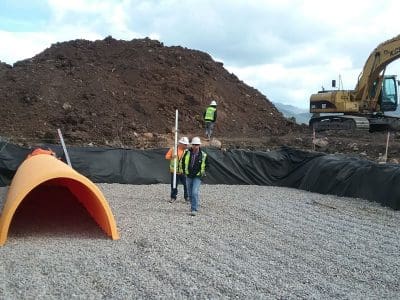 Construction workers in safety gear stand on gravel near an orange pipe, with an excavator and large dirt pile in the background at a construction site.