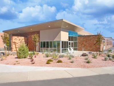 Modern single-story building with large glass windows and brick walls, surrounded by desert landscaping with rocks and drought-tolerant plants under a partly cloudy sky.