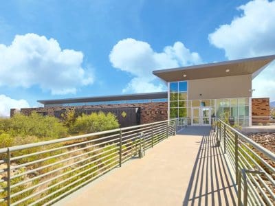 Modern building with large windows and a sloped roof, viewed from a walkway with metal railings under a bright, partly cloudy sky.
