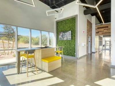 Modern office space with large windows, high ceilings, a tall table with yellow stools, a green plant wall, and polished concrete floors.